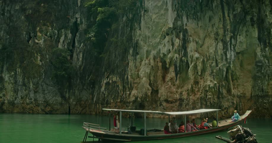 Tourists on a traditional longtail boat float on a calm green lake as the camera reveals the immense limestone cliffs of a national park