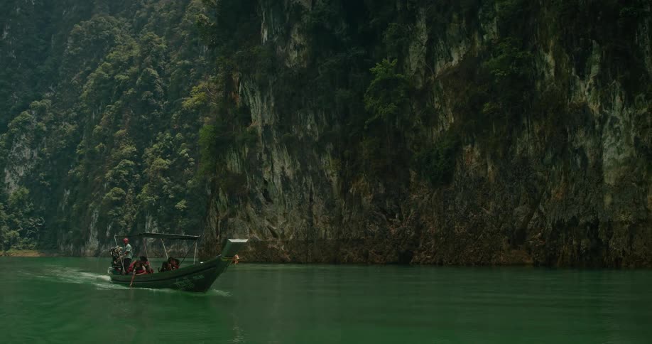 Tourists on a traditional long-tail boat cruise through a stunning green lake surrounded by lush jungle cliffs in Thailand