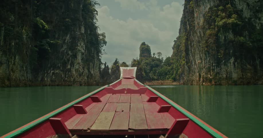 First-person view from a red wooden boat sailing on a tranquil lake through a stunning canyon of jungle-covered cliffs