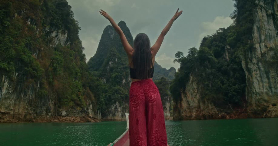 A female traveler raises her arms in freedom and awe while standing on the front of a boat, gliding across an emerald lake through a stunning mountain landscape