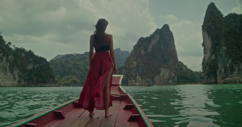 A woman in flowing red pants stands on the bow of a traditional long-tail boat, admiring the majestic mountain scenery of a tranquil lake