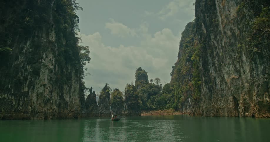 A traditional long-tail boat glides across a tranquil green lake, dwarfed by the majestic limestone cliffs of a remote tropical paradise