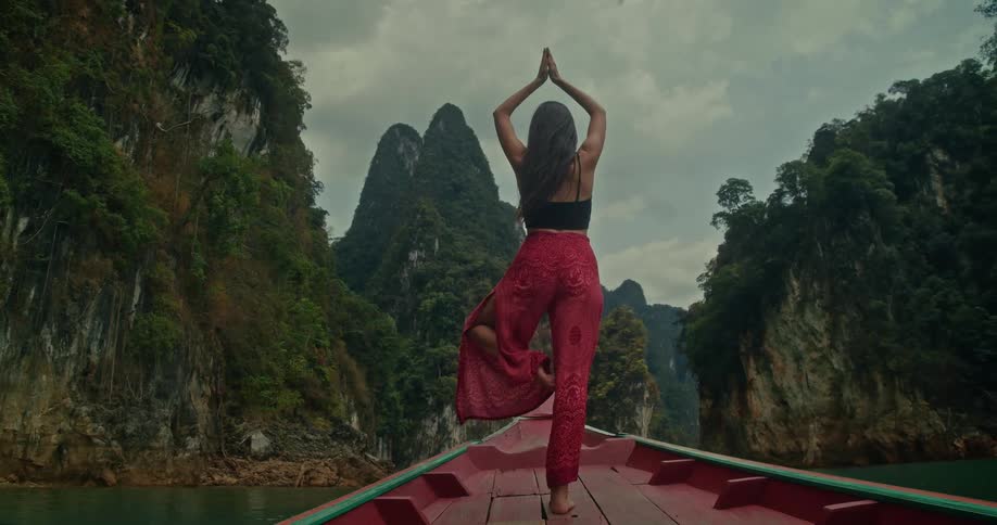 A woman gracefully holds a yoga tree pose on the bow of a traditional boat as it sails through a stunning mountain lake