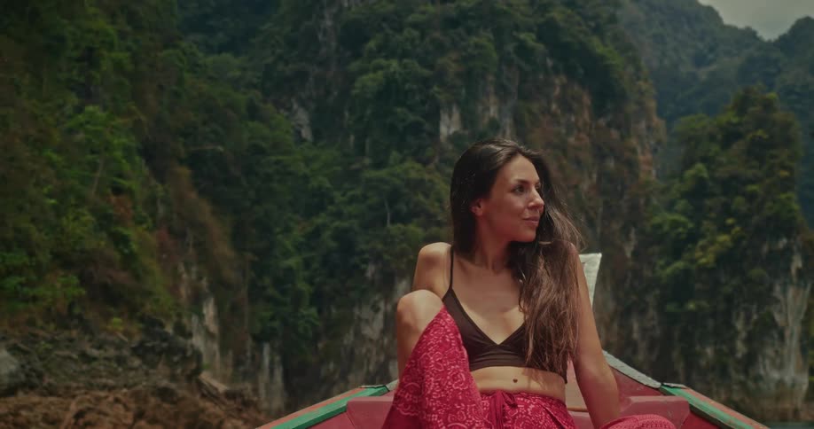 A happy young woman with long dark hair relaxes on the bow of a boat, enjoying the sunshine and lush mountain scenery on a tropical lake