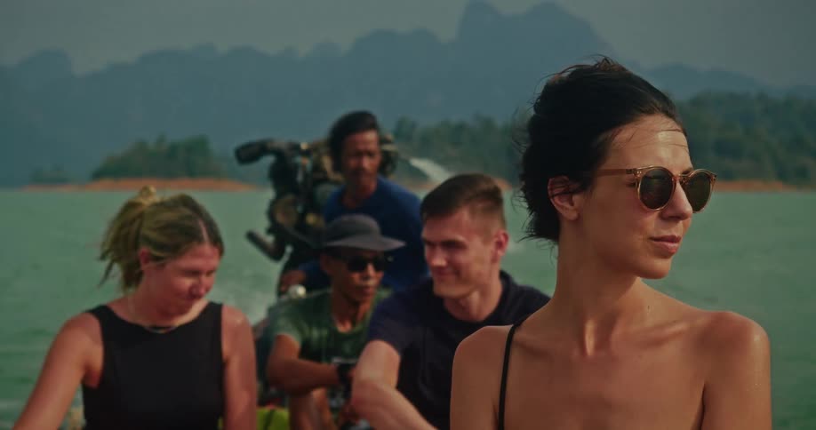 A beautiful young woman with sunglasses enjoys a slow-motion boat tour on a stunning tropical lake with friends