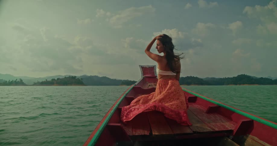 A woman in a flowing skirt enjoys a serene slow-motion boat ride across a vast lake with her hair blowing in the wind