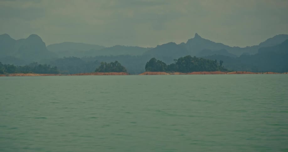 Tilting shot reveals a vast, tranquil lake surrounded by hazy, lush green mountains and islands on an overcast day