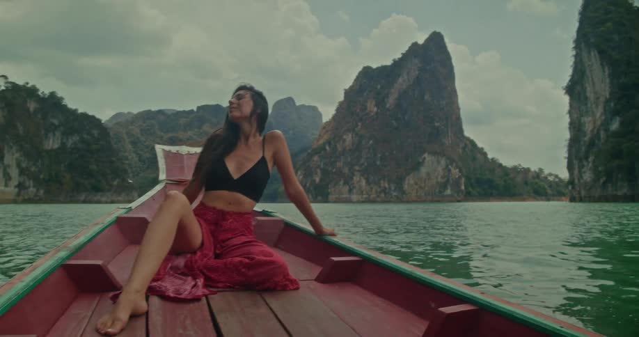 A woman relaxes on the bow of a traditional wooden boat, sailing across a lake surrounded by majestic mountains in Thailand