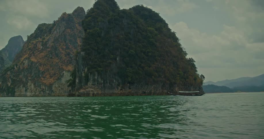 A traditional long-tail boat cruises across a vast lake surrounded by massive limestone cliffs on an overcast day in Thailand
