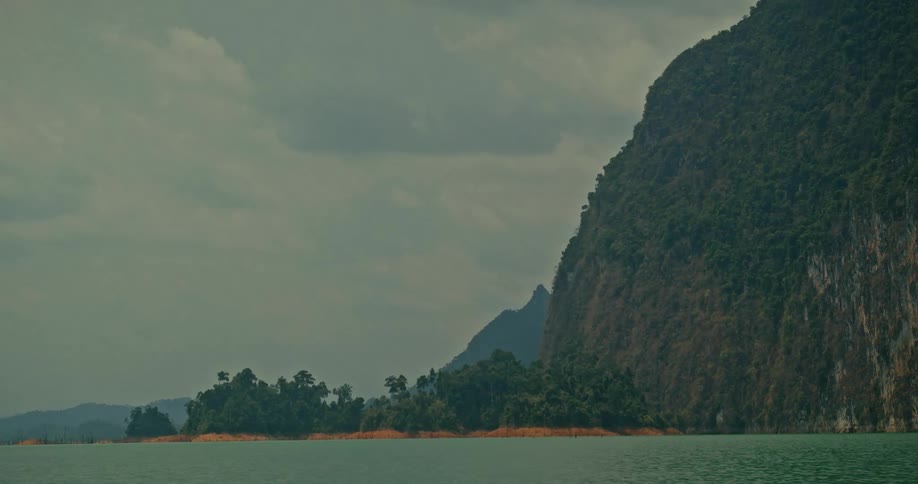 A tranquil wide shot of a calm lake and steep, forested mountain under an overcast sky