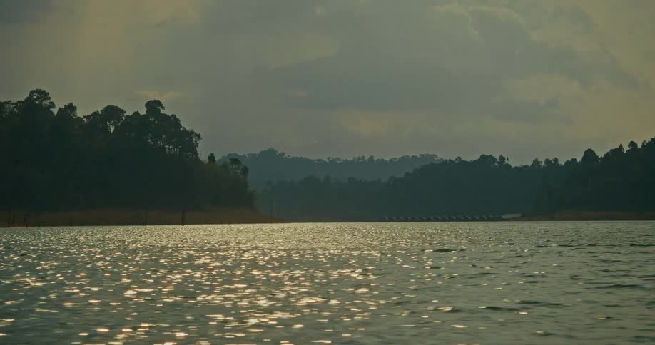 The sun glitters on the surface of a vast lake surrounded by a tropical rainforest under a cloudy sky