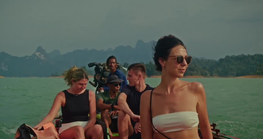 A stylish young woman with windswept hair enjoys a slow-motion boat tour on a tranquil lake surrounded by majestic mountains