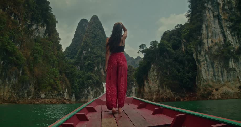 A woman stands at the bow of a traditional boat, sailing through a majestic canyon of towering limestone cliffs on a serene lake