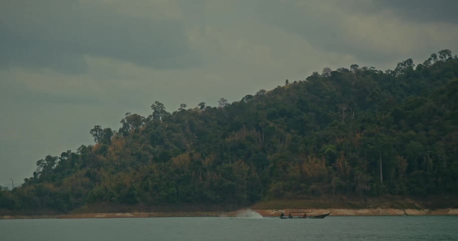 A traditional longtail boat speeds across a vast lake with a dense, tree-covered mountain in the background on a cloudy day