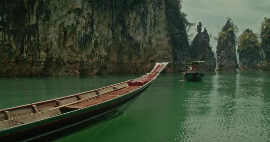 A traditional long-tail boat floats peacefully on an emerald lake, with a slow zoom revealing towering limestone karsts