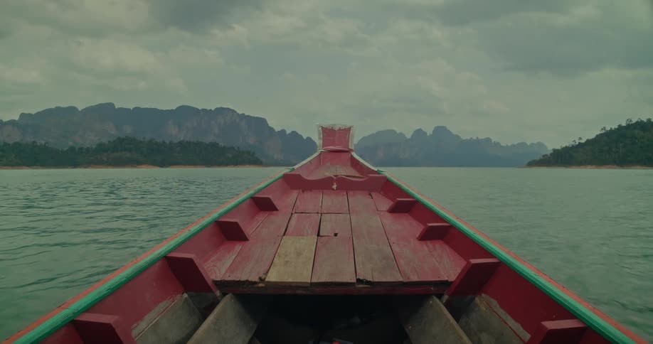 A serene point-of-view shot from a traditional red long-tail boat cruising across a tranquil lake toward majestic mountains on a cloudy day