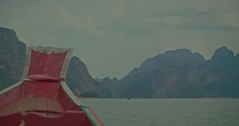 View from the bow of a traditional red longtail boat sailing across a serene lake surrounded by majestic limestone mountains on a cloudy day