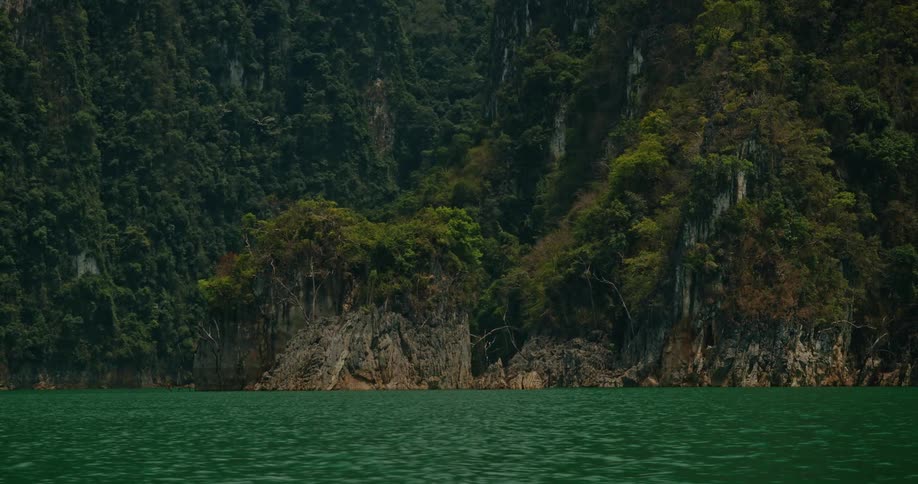 A slow zoom out from a narrow mountain pass reveals the stunning landscape of lush green cliffs rising from a tranquil emerald lake