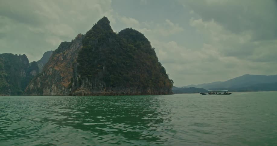A tour boat cruises across the calm green water beside a large, tree-covered karst mountain under a cloudy sky