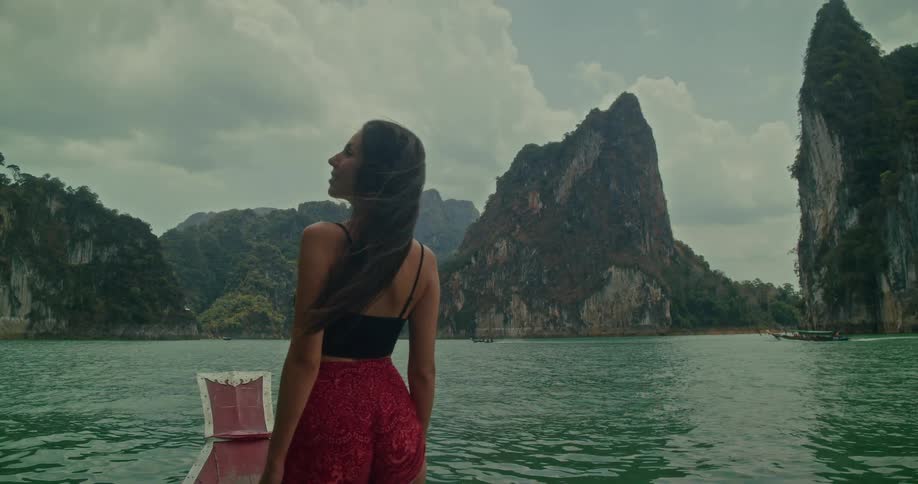 A woman with long flowing hair stands at the bow of a boat, enjoying the view of the vast lake and majestic mountains in slow motion