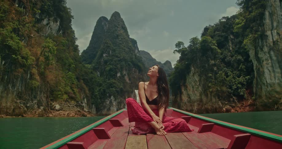 A blissful young woman soaks up the sun while cruising on a long-tail boat through a spectacular tropical bay with towering green cliffs