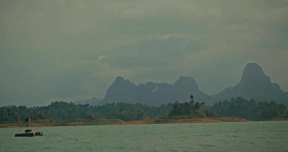A serene lake with a floating buoy is slowly revealed to have majestic mountains in the background under an overcast sky