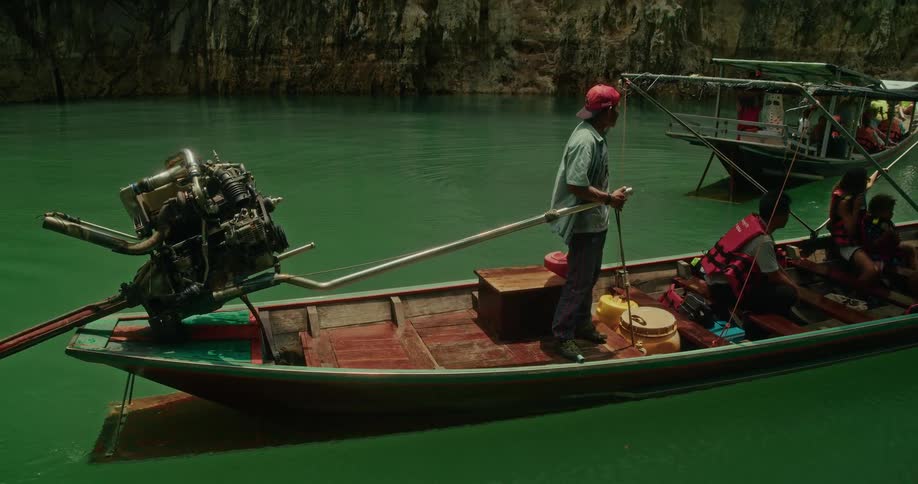 A Thai tour guide pilots a traditional long-tail boat carrying tourists across a stunning emerald green lake