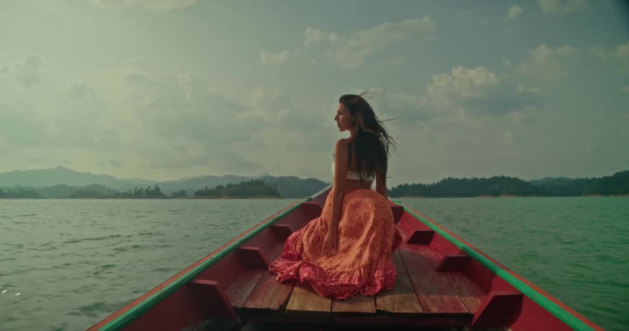 A young woman in a colorful skirt sits on the bow of a boat, enjoying the view of the vast lake and mountains