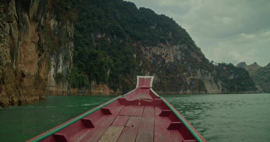 POV from a traditional red long-tail boat gliding through a serene lake towards towering, jungle-covered limestone cliffs