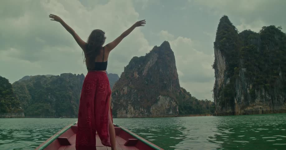 A woman in a flowing red skirt stands on the bow of a traditional boat, raising her arms in freedom while sailing across a lake surrounded by majestic limestone cliffs