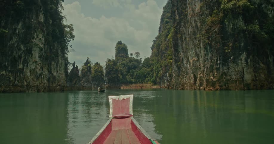 A traditional red wooden boat gently sails through a beautiful, serene lake surrounded by towering jungle-clad limestone cliffs in Thailand