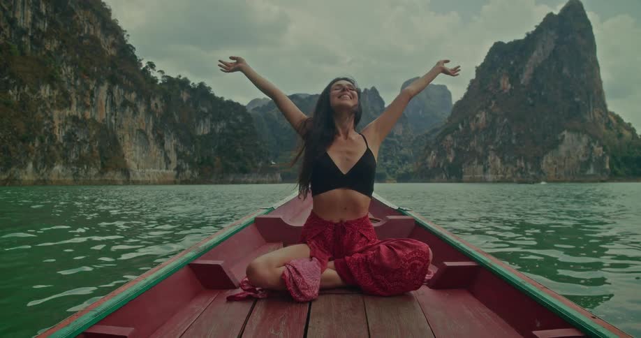 A beautiful woman traveler enjoys a peaceful longtail boat ride on a lake, raising her arms in happiness against a stunning backdrop of majestic limestone mountains