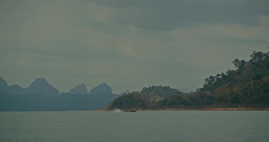 A long-tail boat travels across a vast lake against a stunning backdrop of misty mountains on an overcast day