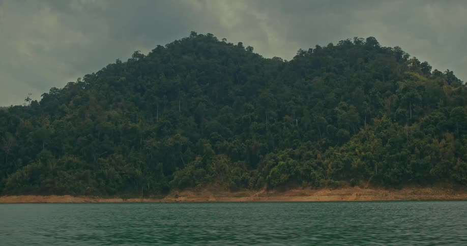 A calm, rippling lake sits before a dense, green forest-covered hill on a cloudy day