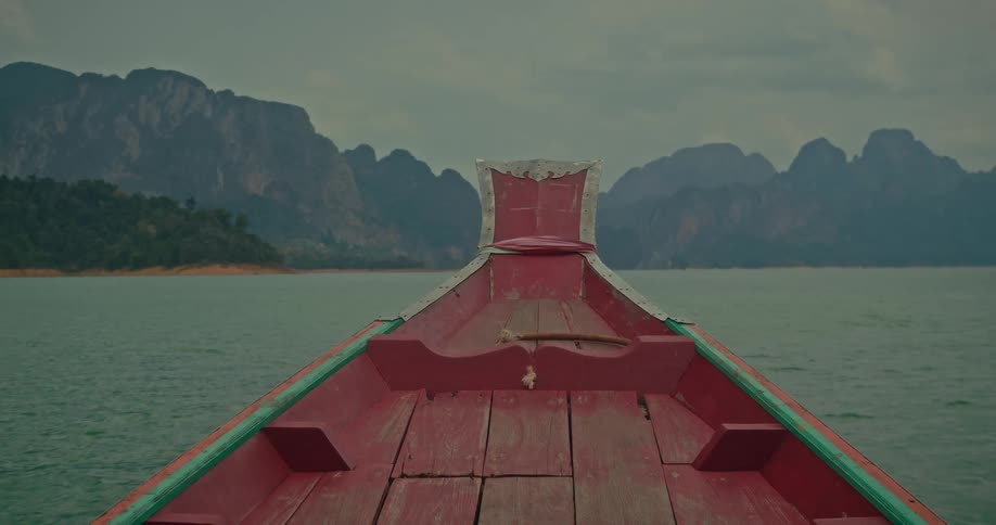 A point-of-view shot from the front of a rustic red boat moving across a tranquil lake towards stunning mountain karsts under a cloudy sky