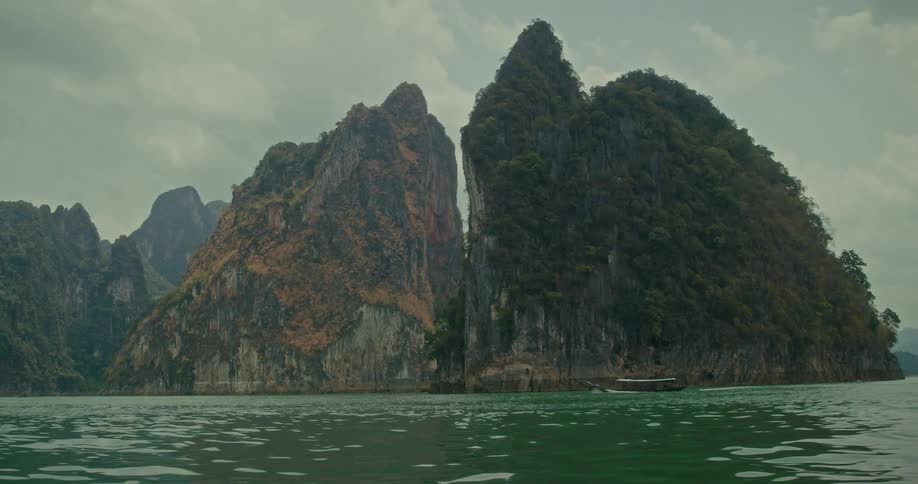 A long-tail boat sails across a tranquil lake past a dramatic split limestone mountain in Khao Sok National Park, Thailand