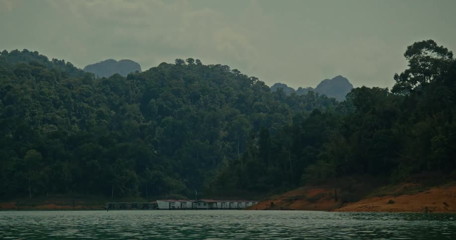 Floating houses sit peacefully on a tranquil lake at the base of a dense rainforest mountain range on an overcast day