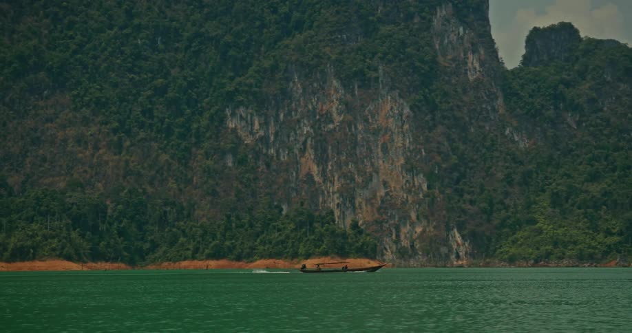 A traditional long-tail boat cruises across a vast emerald lake, set against a stunning backdrop of lush, jungle-covered mountains