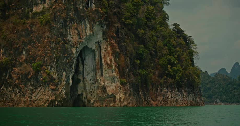 A sheer limestone cliff with a mysterious cave entrance, covered in jungle foliage, rises from the tranquil green waters of a lake