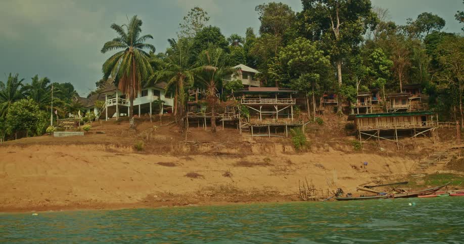 A peaceful tropical village with rustic bungalows sits on the sandy bank of a calm river surrounded by lush jungle