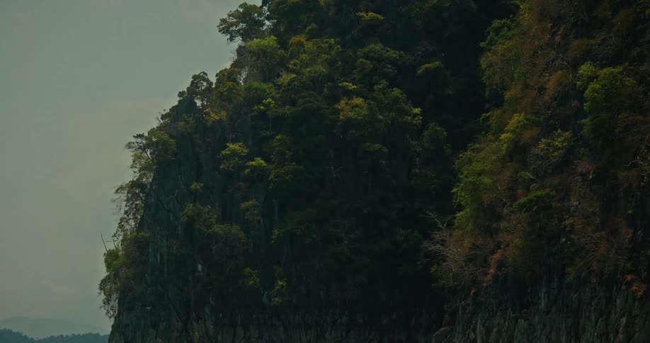A slow zoom out reveals a towering limestone cliff covered in lush tropical jungle rising from the calm, emerald waters of a serene lake