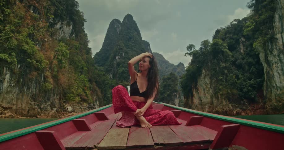 A serene woman on the bow of a traditional long-tail boat drifts through a stunning tropical lake, surrounded by towering jungle-covered mountains