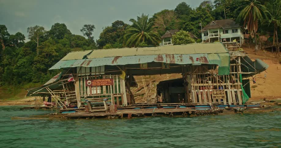 A rustic and dilapidated floating raft house bobs on the water's surface next to a lush, forested shoreline