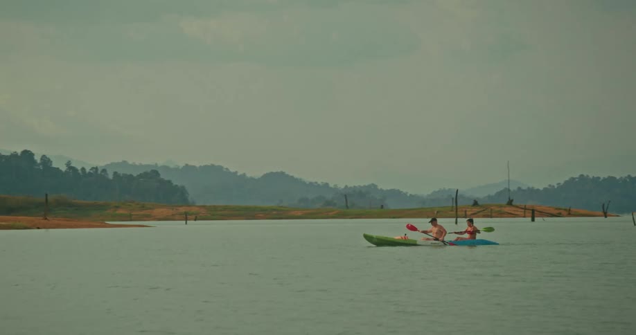 A wide shot follows two men as they kayak together across a vast, calm lake surrounded by misty, forested mountains