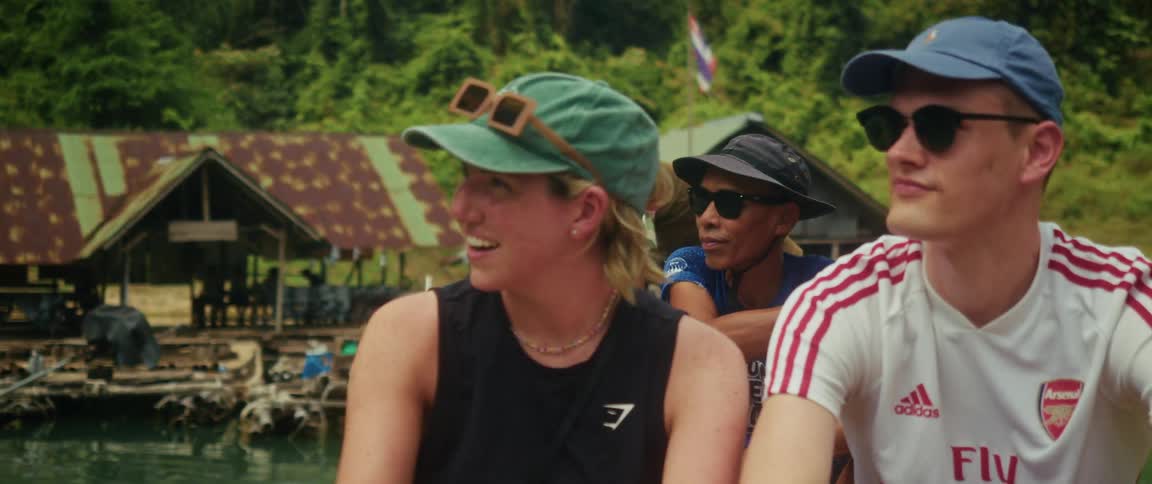 A young woman wearing a green cap smiles and waves while on a boat tour with friends through a riverside village in Thailand
