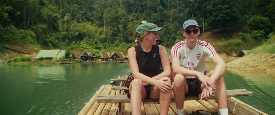 A young man and woman sit together on a bamboo raft as it floats across a tranquil jungle lake