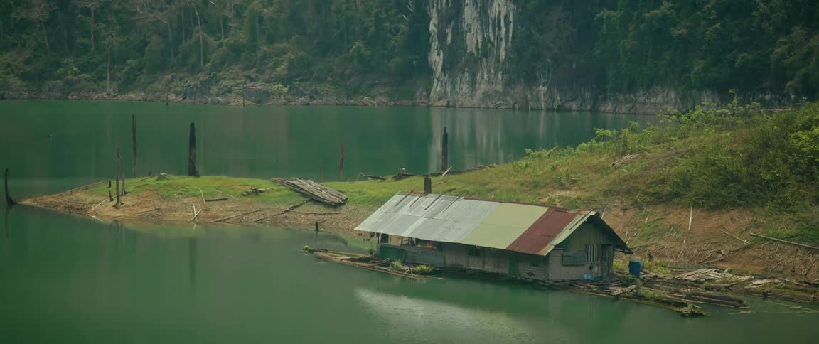 A rustic floating house sits on the calm green waters of a remote lake at the base of a lush jungle cliff