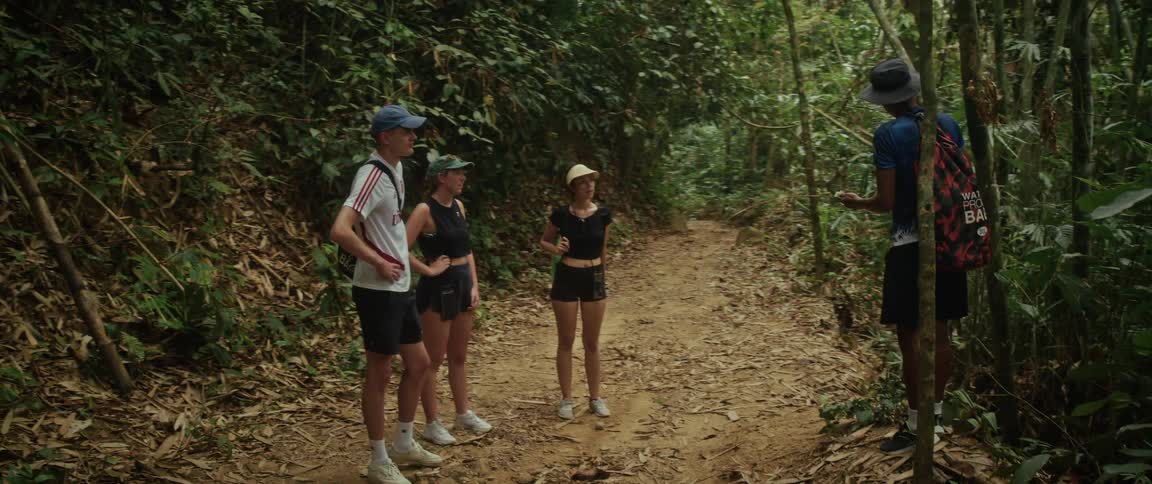 A local guide shares knowledge with a group of young hikers on a dirt trail in a dense tropical jungle
