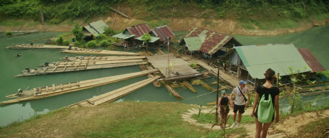 A group of tourists look down from a grassy hill at a floating village with traditional bamboo rafts docked on a river in Thailand