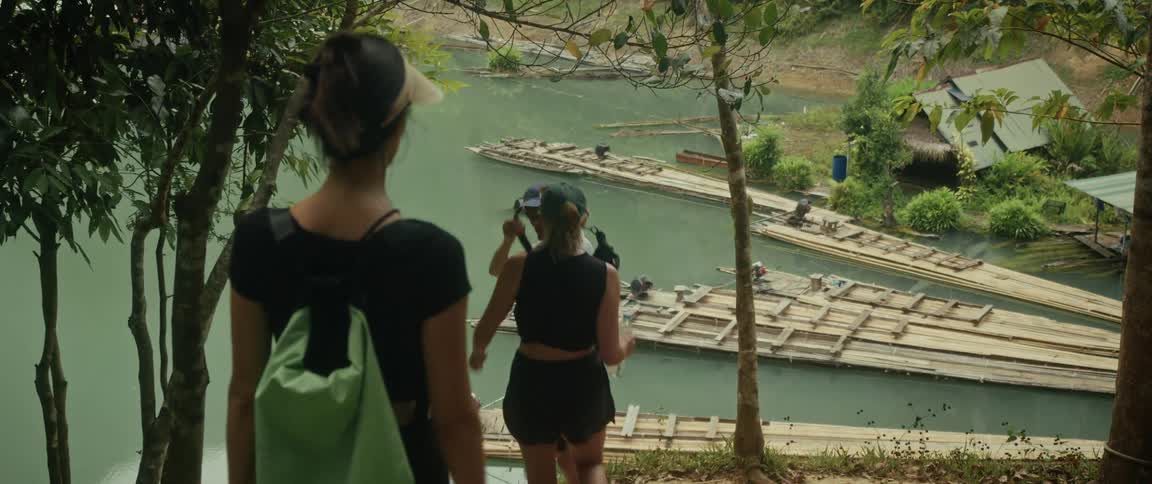 A group of tourists hike down a path in a lush jungle toward bamboo rafts on a river
